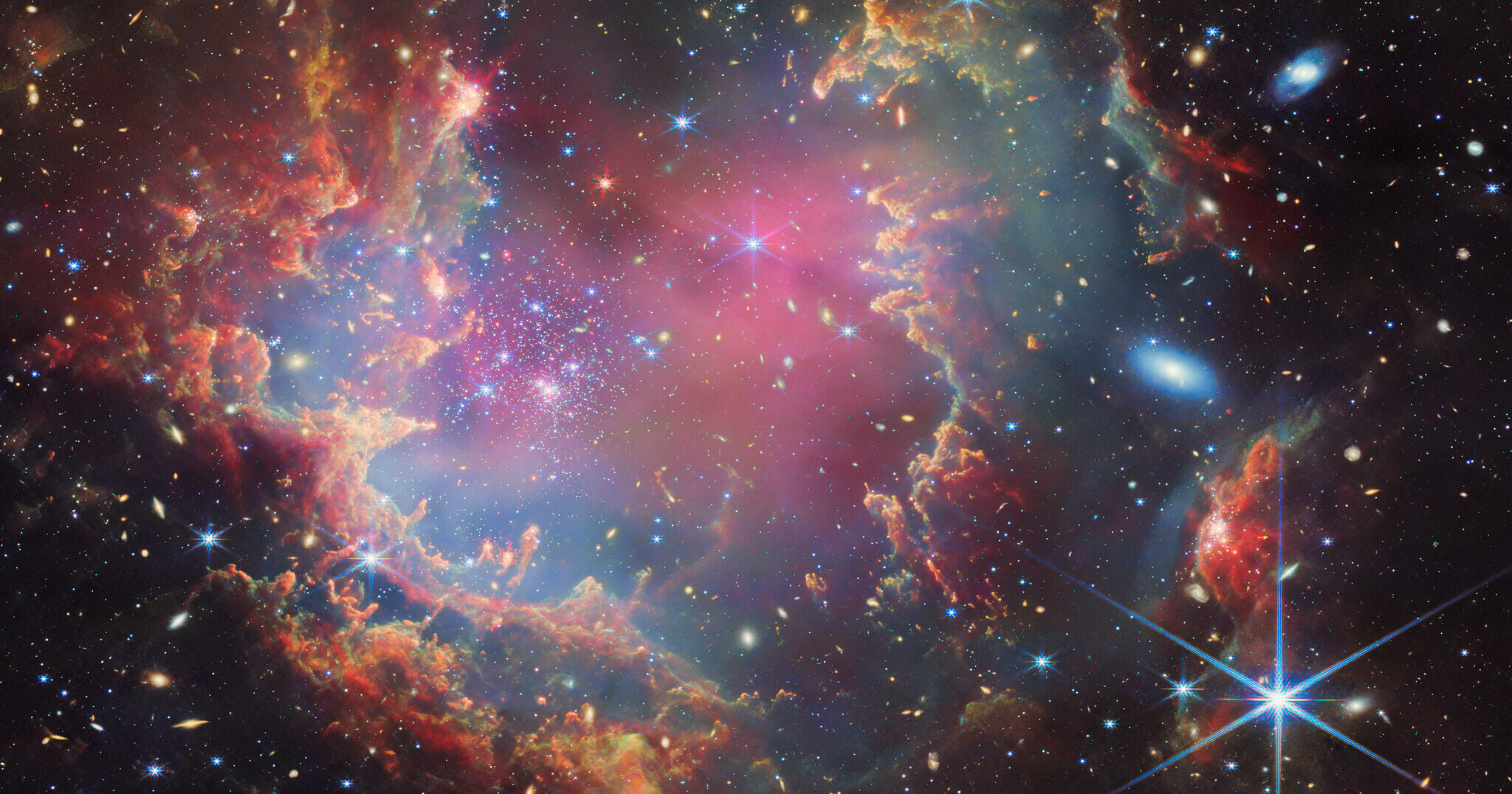 A star cluster is shown inside a large nebula of many-coloured gas and dust. The material forms dark ridges and peaks of gas and dust surrounding the cluster, lit on the inner side, while layers of diffuse, translucent clouds blanket over them. Around and within the gas, a huge number of distant galaxies can be seen, some quite large, as well as a few stars nearer to us which are very large and bright. Credit: ESA/Webb, NASA & CSA, P. Zeidler, E. Sabbi, A. Nota, M. Zamani (ESA/Webb)