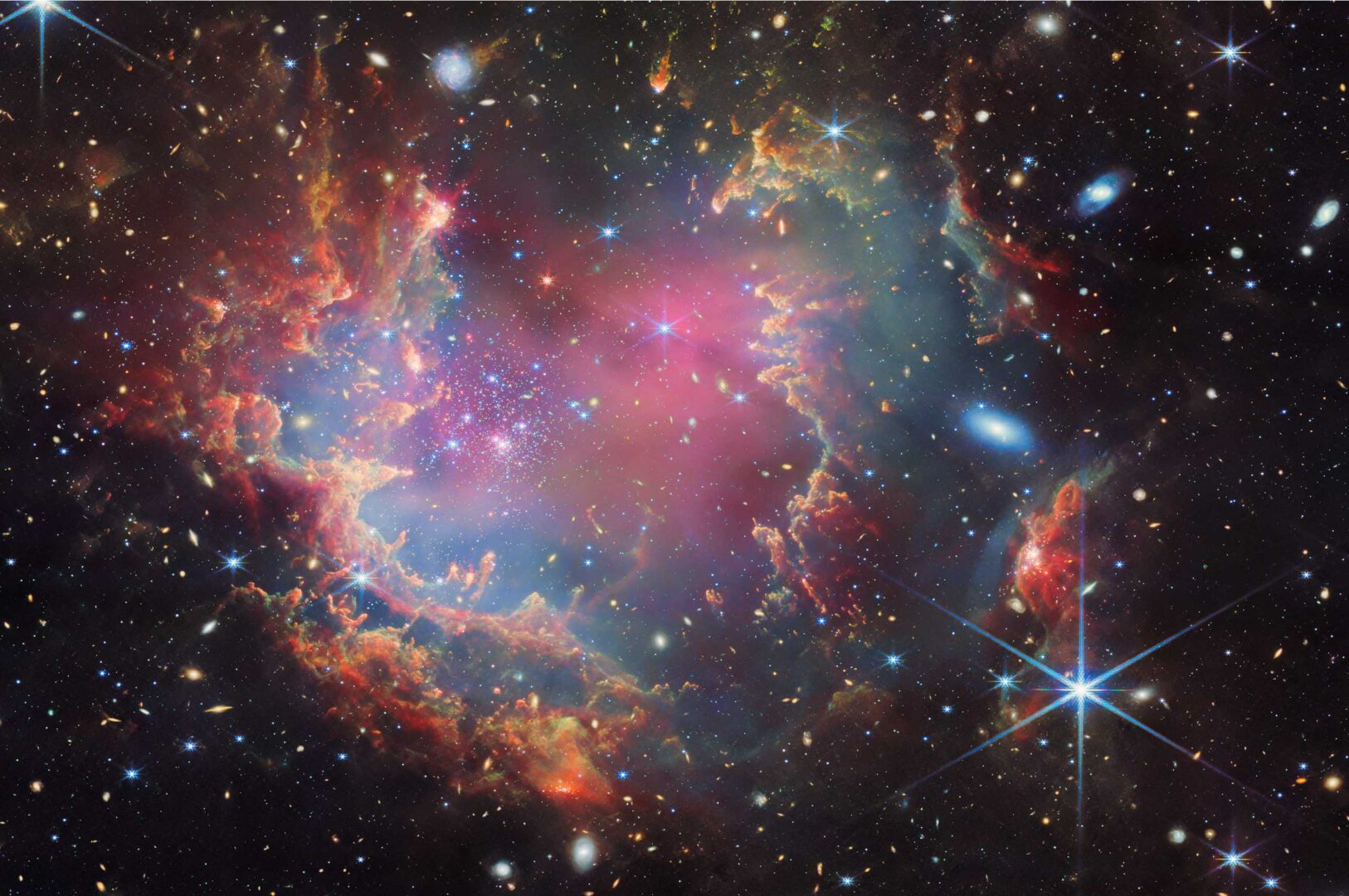 A star cluster is shown inside a large nebula of many-coloured gas and dust. The material forms dark ridges and peaks of gas and dust surrounding the cluster, lit on the inner side, while layers of diffuse, translucent clouds blanket over them. Around and within the gas, a huge number of distant galaxies can be seen, some quite large, as well as a few stars nearer to us which are very large and bright. Credit: ESA/Webb, NASA & CSA, P. Zeidler, E. Sabbi, A. Nota, M. Zamani (ESA/Webb)